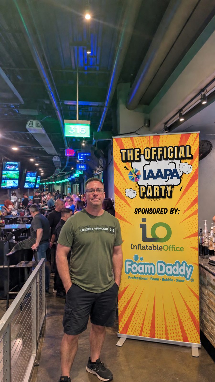 Guest standing beside the tall yellow Official IAAPA Party banner sponsored by Inflatable Office and Foam Daddy in the Topgolf bays